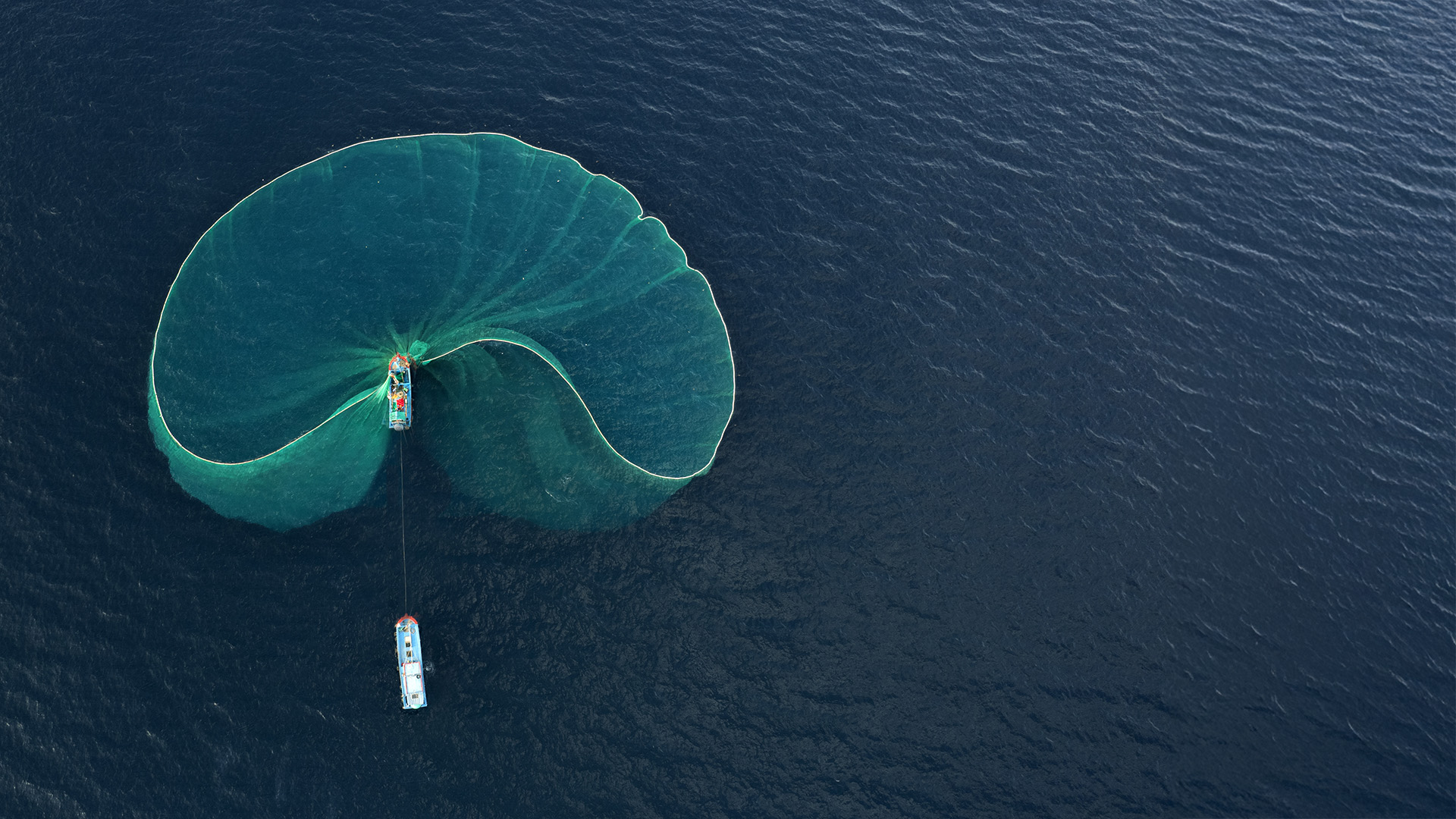 Aerial view of a lone fishing boat with its net deployed over a dark sea.