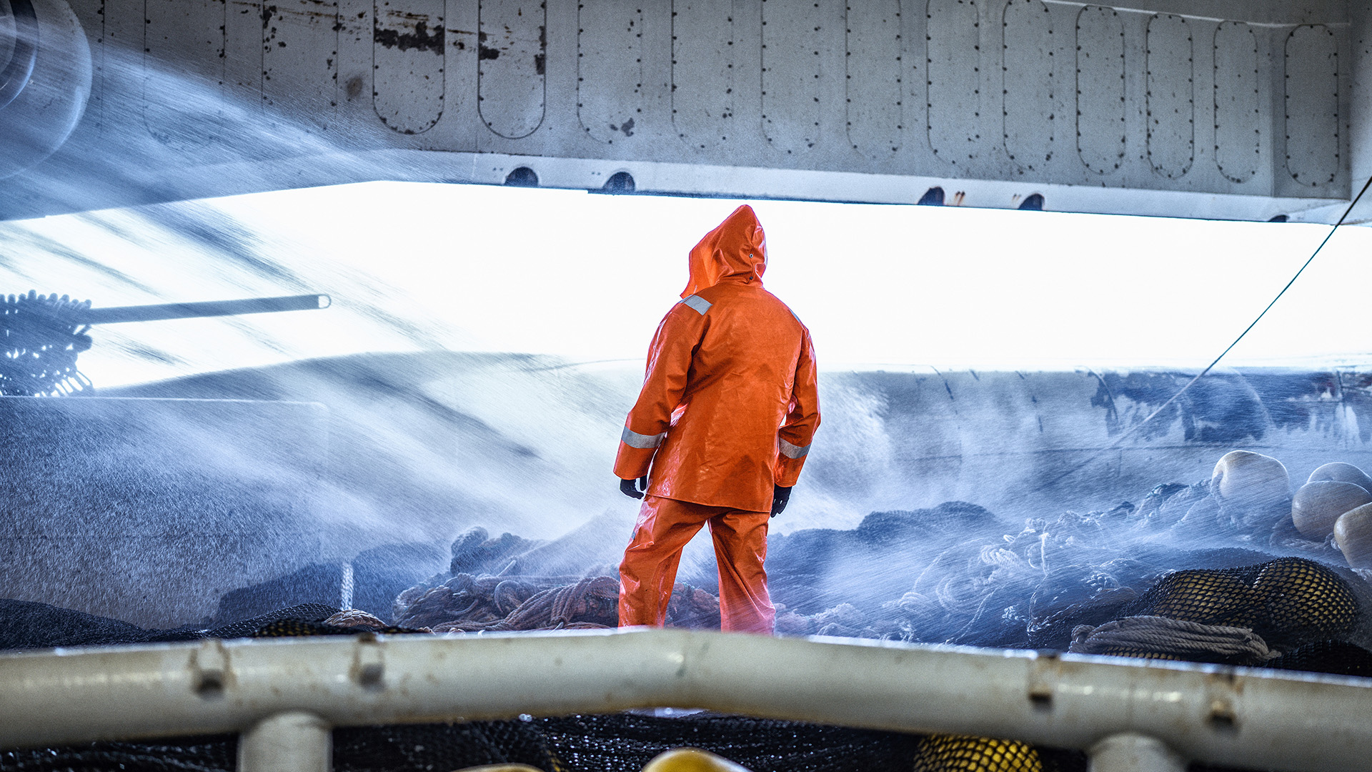 Ocean spray soaks a fisherman in a bright orange jacket and pants, as he stands on the deck of a commercial fishing boat