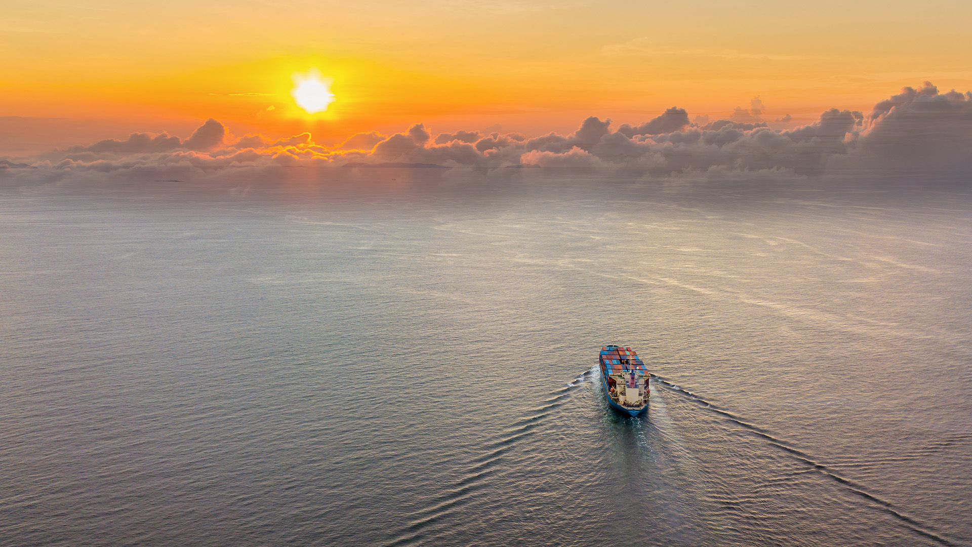 A container ship heads off across grey ocean waters toward an orange sunset