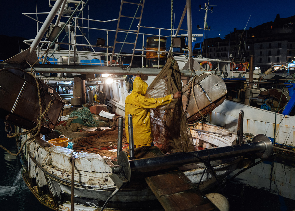 A fisherman in wet coveralls works on the cluttered deck of an old ship at night