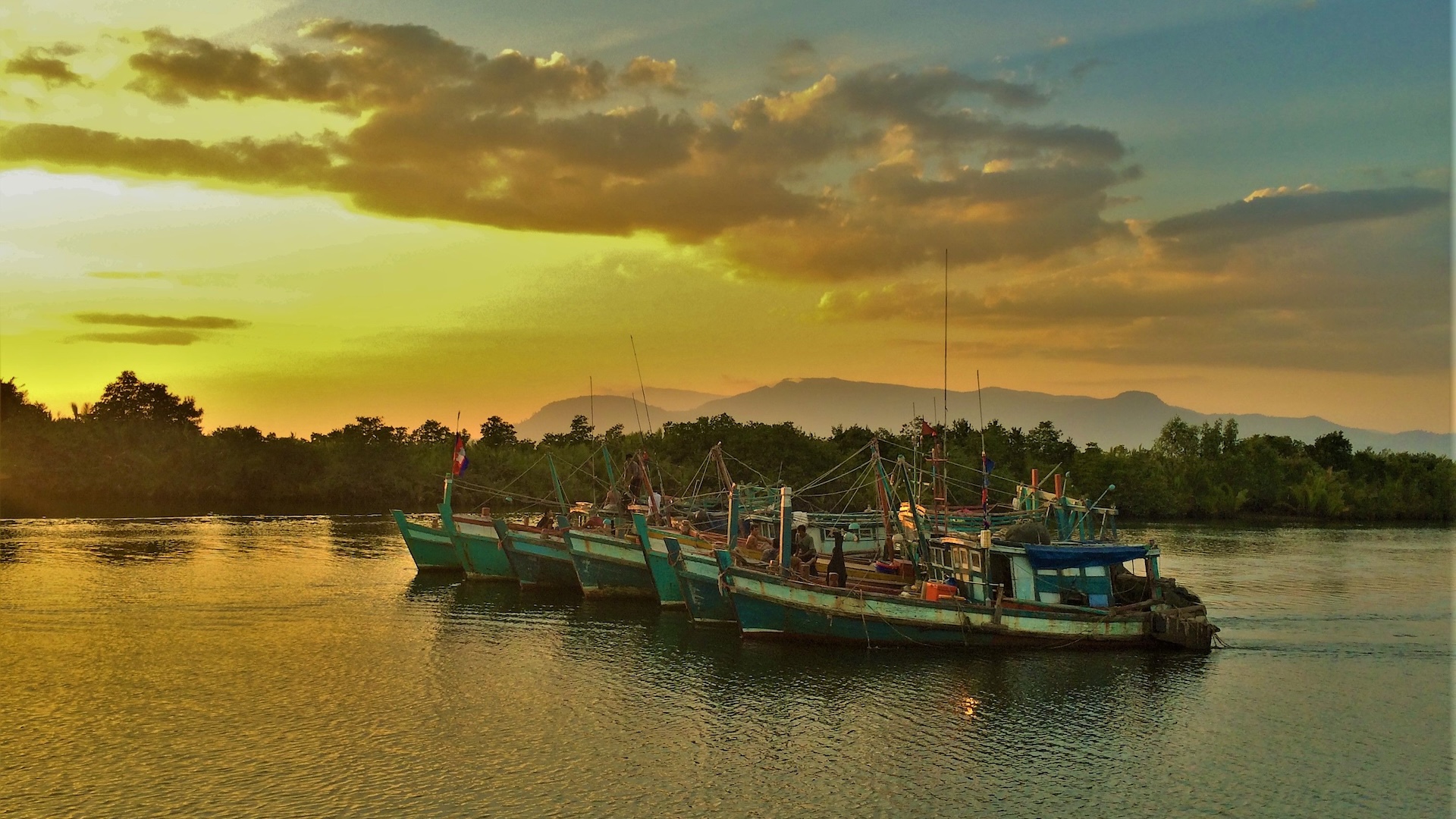Fishing boats on the water in Cambodia during a vibrant sunset with mountains in the background.