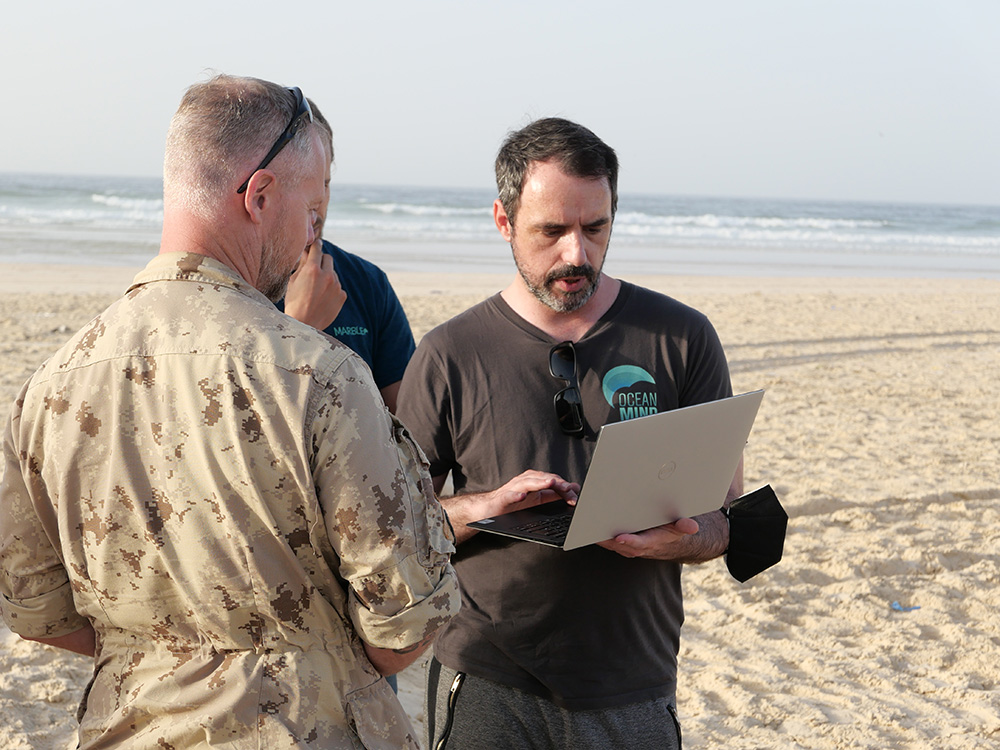 An OceanMind team member consults with an enforcement official reviewing a handheld laptop computer at the water's edge